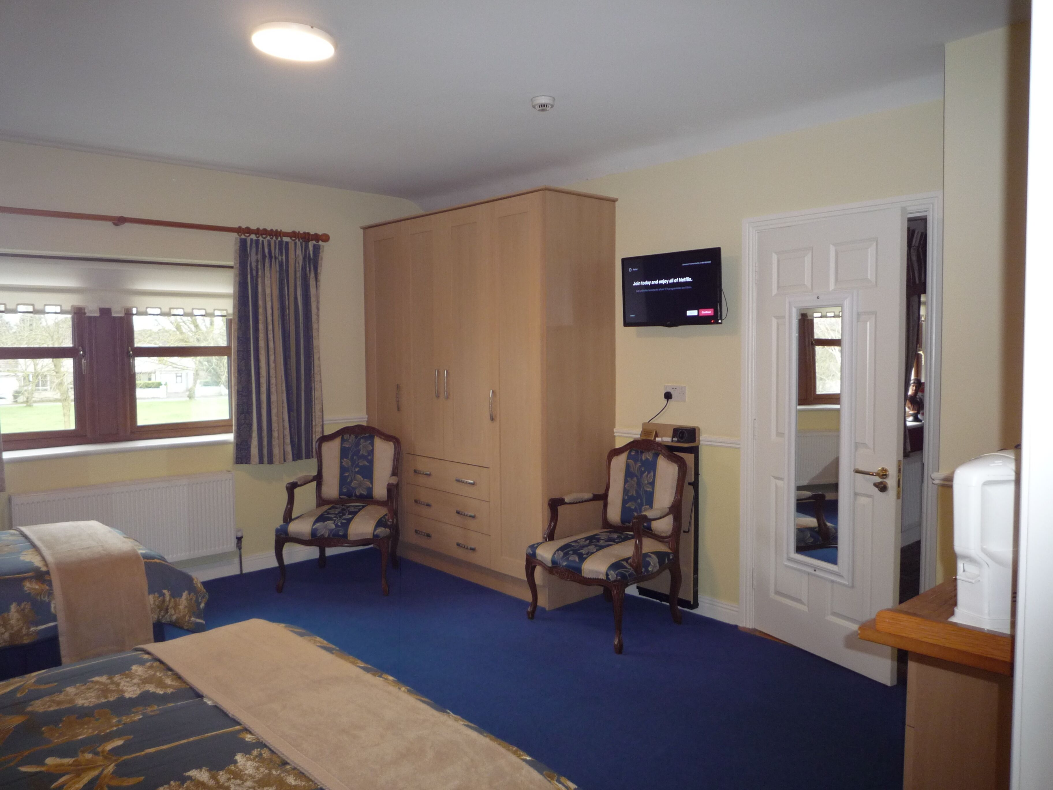 A bedroom with a bed, two chairs, a wardrobe, a television, and a door.