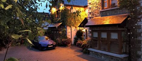 A stone house with a car parked in front, surrounded by greenery and a brick driveway.