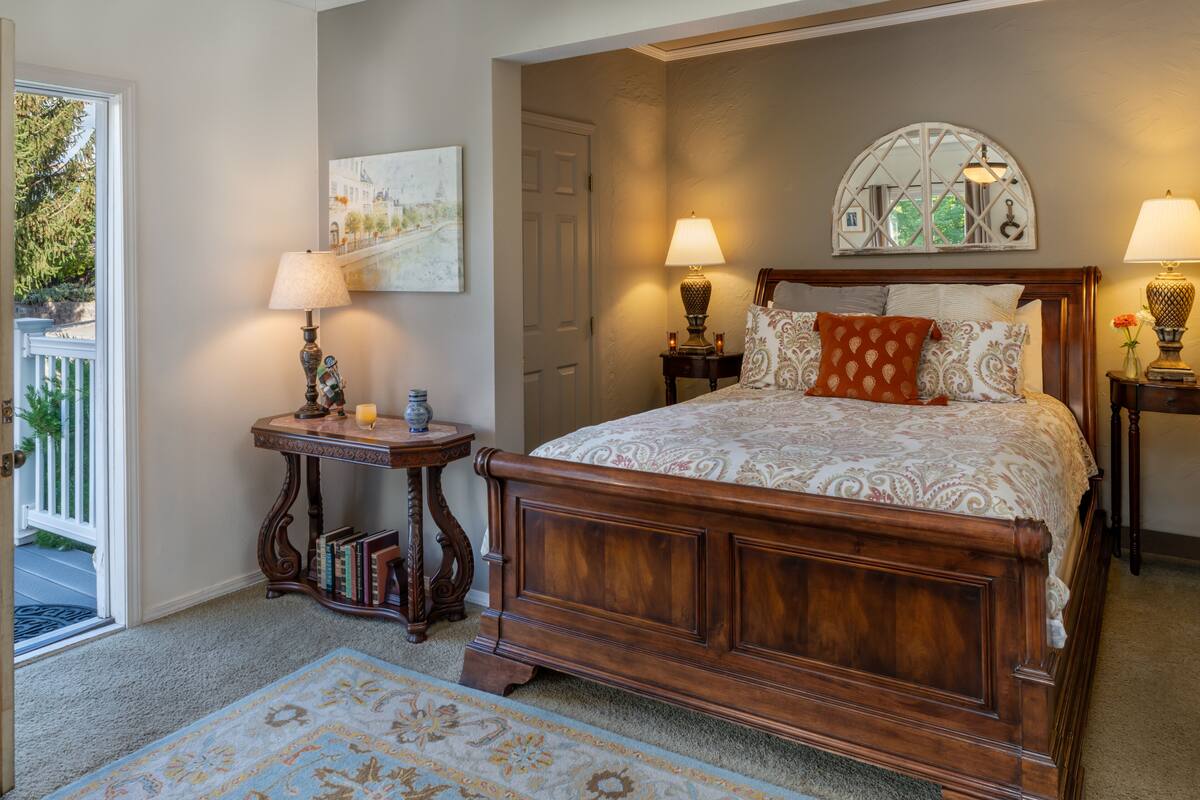 Um quarto com cama de madeira, duas mesas de cabeceira com luminárias, um tapete, uma porta e um quadro na parede.