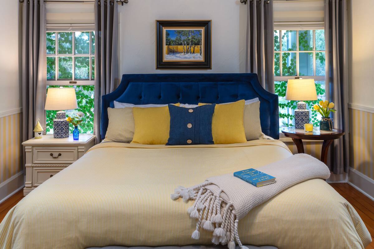 Cama bem arrumada com cabeceira azul, almofadas amarelas e azuis, e um livro sobre a cama. Há duas mesas de cabeceira com abajures e objetos decorativos.