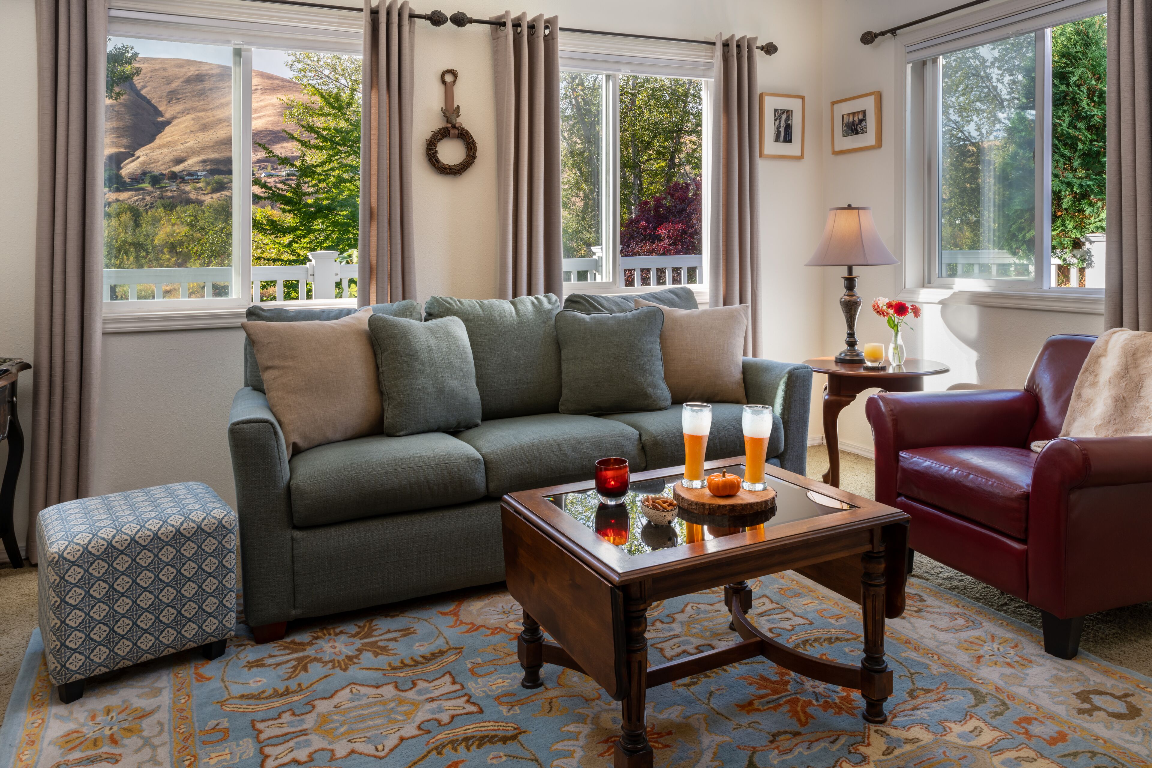 A cozy living room with a sofa, ottoman, and coffee table, a red armchair, and a view of a mountainous landscape through large windows.
