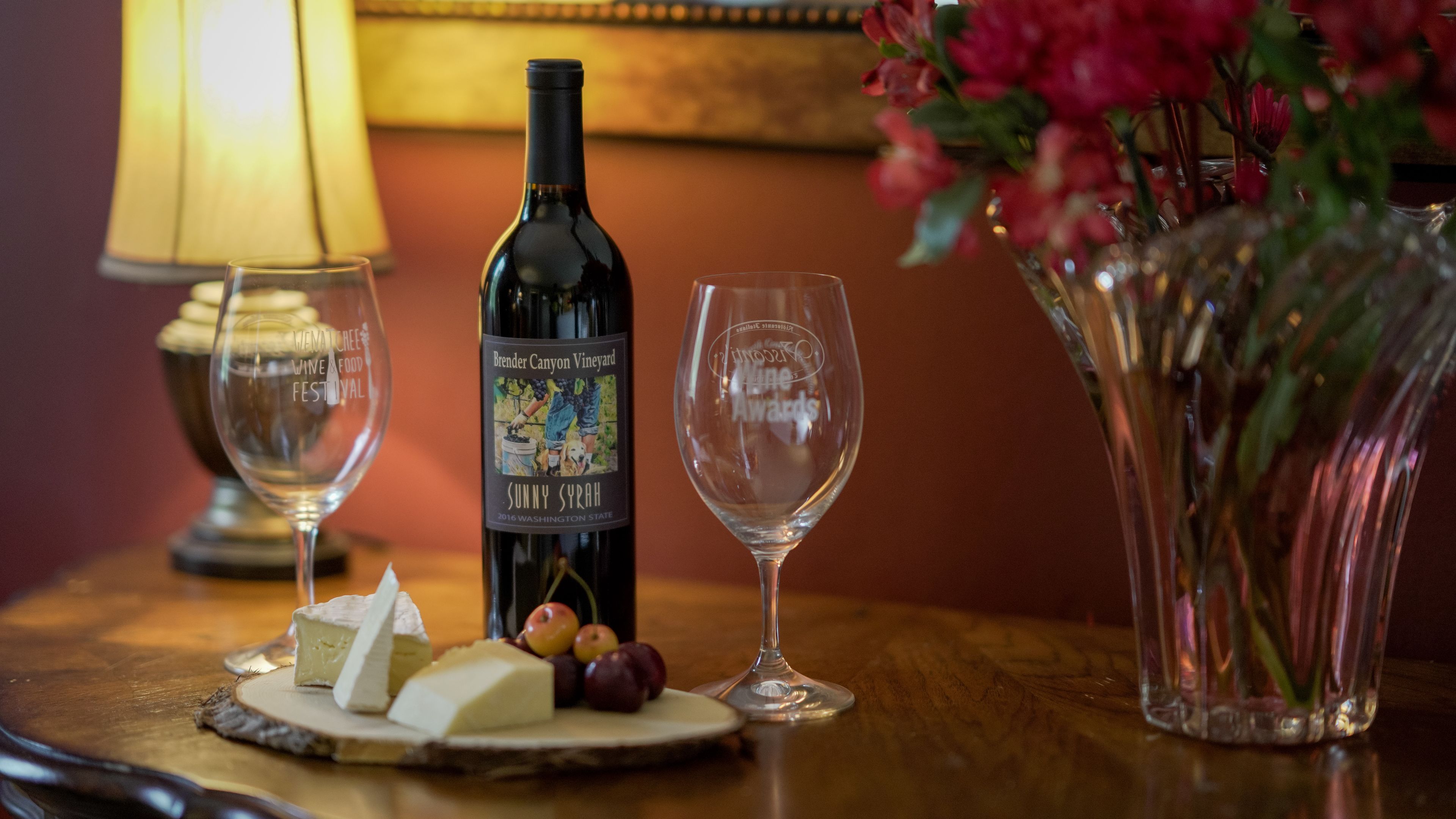 A bottle of Brander Canyon Vineyard Sunny Syrah wine, a glass, and a plate with cheese and grapes on a wooden table.