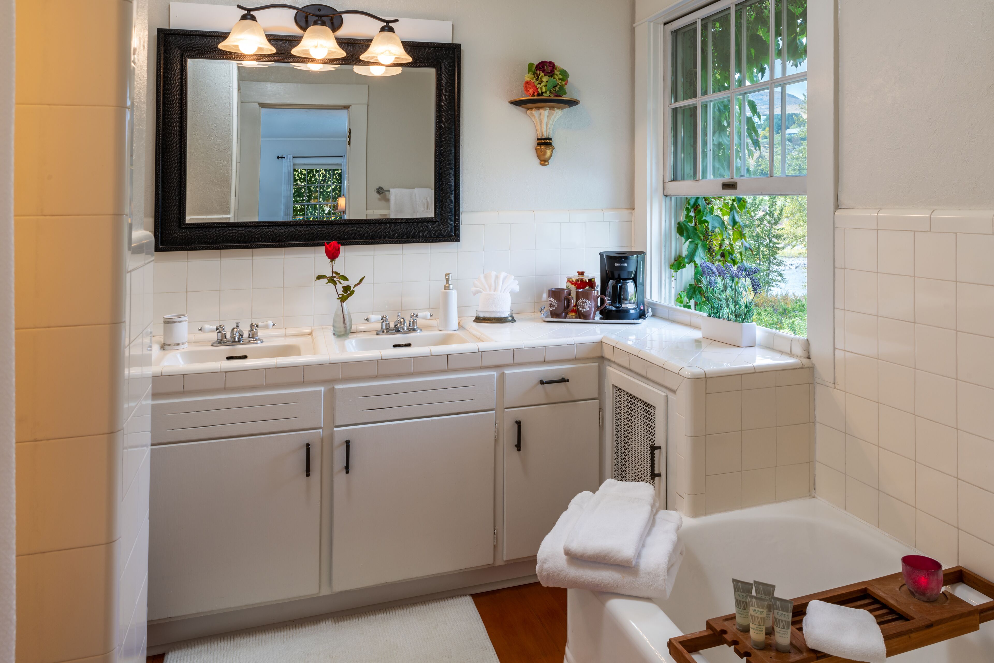 A modern bathroom with a large mirror, white tiles, a window with a view, and a wooden tray with toiletries.