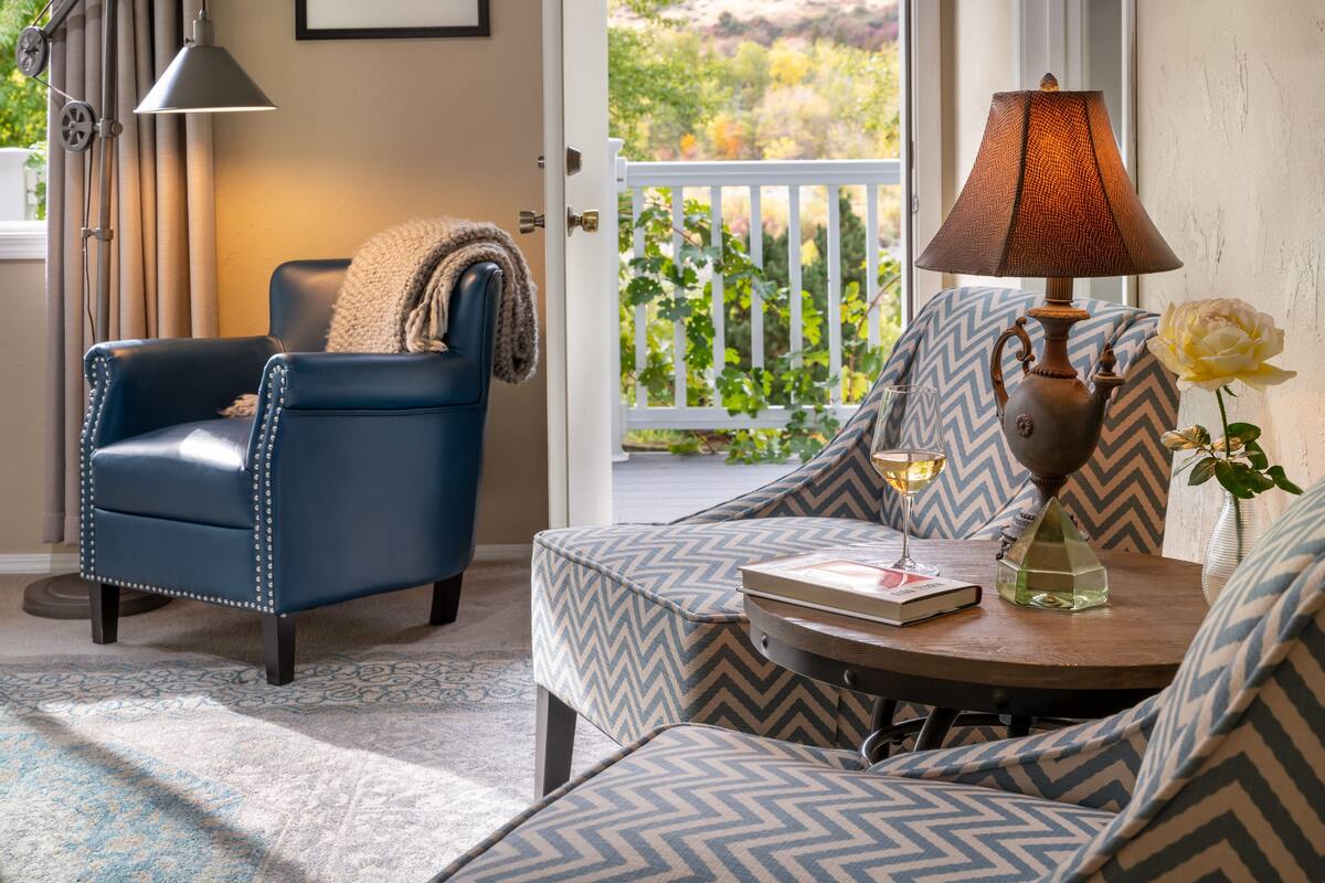Quarto com poltrona azul, sofá estampado, mesa pequena com abajur, vaso com rosa e vista para o verde através de uma porta.