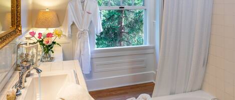 A bathroom with a sink, mirror, and bathtub. There is a window with a view of trees, a towel rack, and a candle on the sink.
