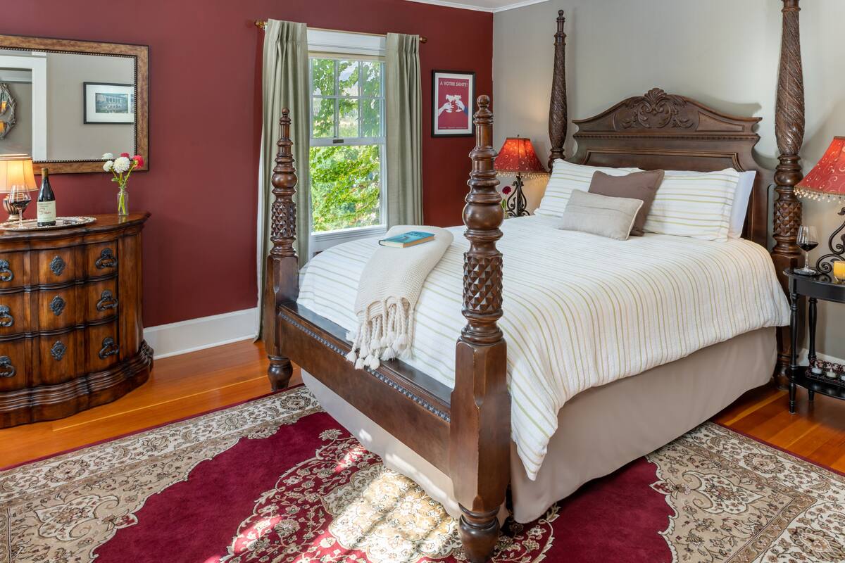 Uma cama com dossel e cabeceira de madeira, uma mesa de cabeceira com abajur e um tapete com padrão em vermelho e bege.