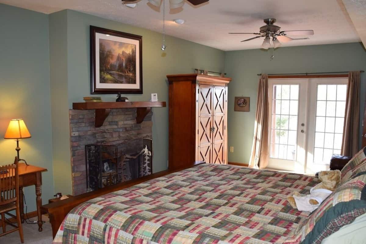 A bedroom with a large bed, a fireplace, a desk with a lamp, and a door with curtains.