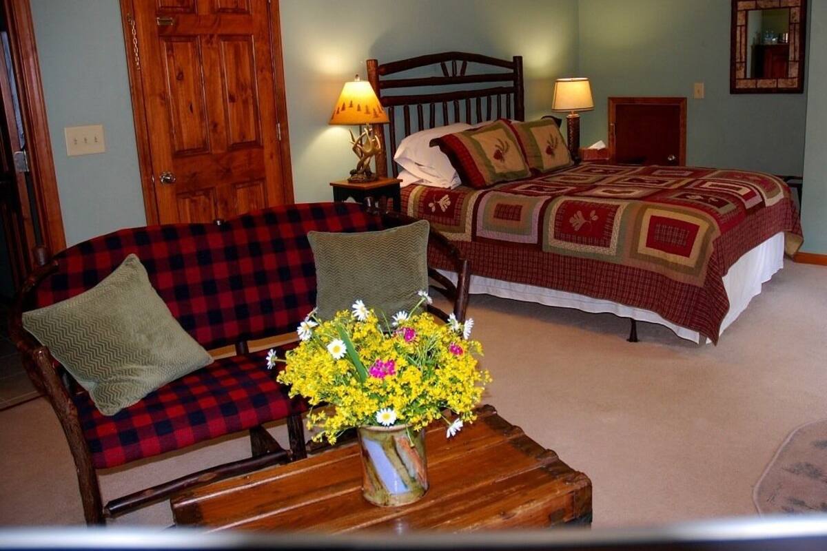 A bedroom with a wooden bed, a red and black checkered sofa, a wooden nightstand with a lamp, and a vase of yellow flowers.