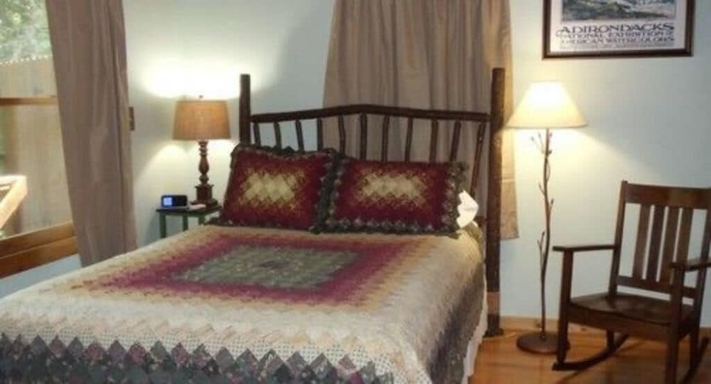 Una cama bien hecha con una colcha estampada, un cabecero de madera, dos lámparas de noche, una silla y un cuadro enmarcado en la pared.