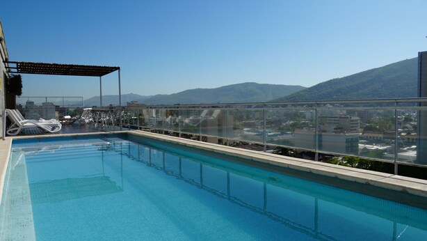 A rooftop pool with clear blue water, lounge chairs, and a glass railing overlooking a cityscape and mountains.