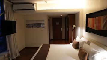 A hotel room with a large bed, a flat-screen TV, an air conditioning unit, a painting on the wall, and a lamp.