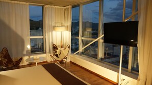 A hotel room with a large window, a flat-screen TV, a lamp, a chair, and a small table.