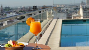 A glass of orange juice with a slice of orange on a table by a pool with a city view.