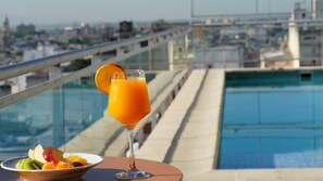 A glass of orange juice with a slice of orange on a table by a pool with a city view.