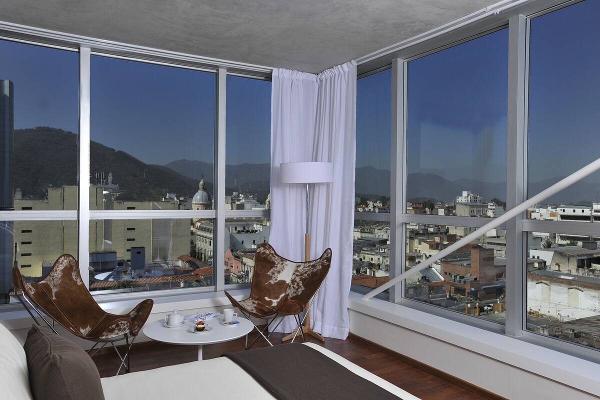A high-rise room with large windows, a sofa, two chairs, a small table with a teapot, and a lamp.