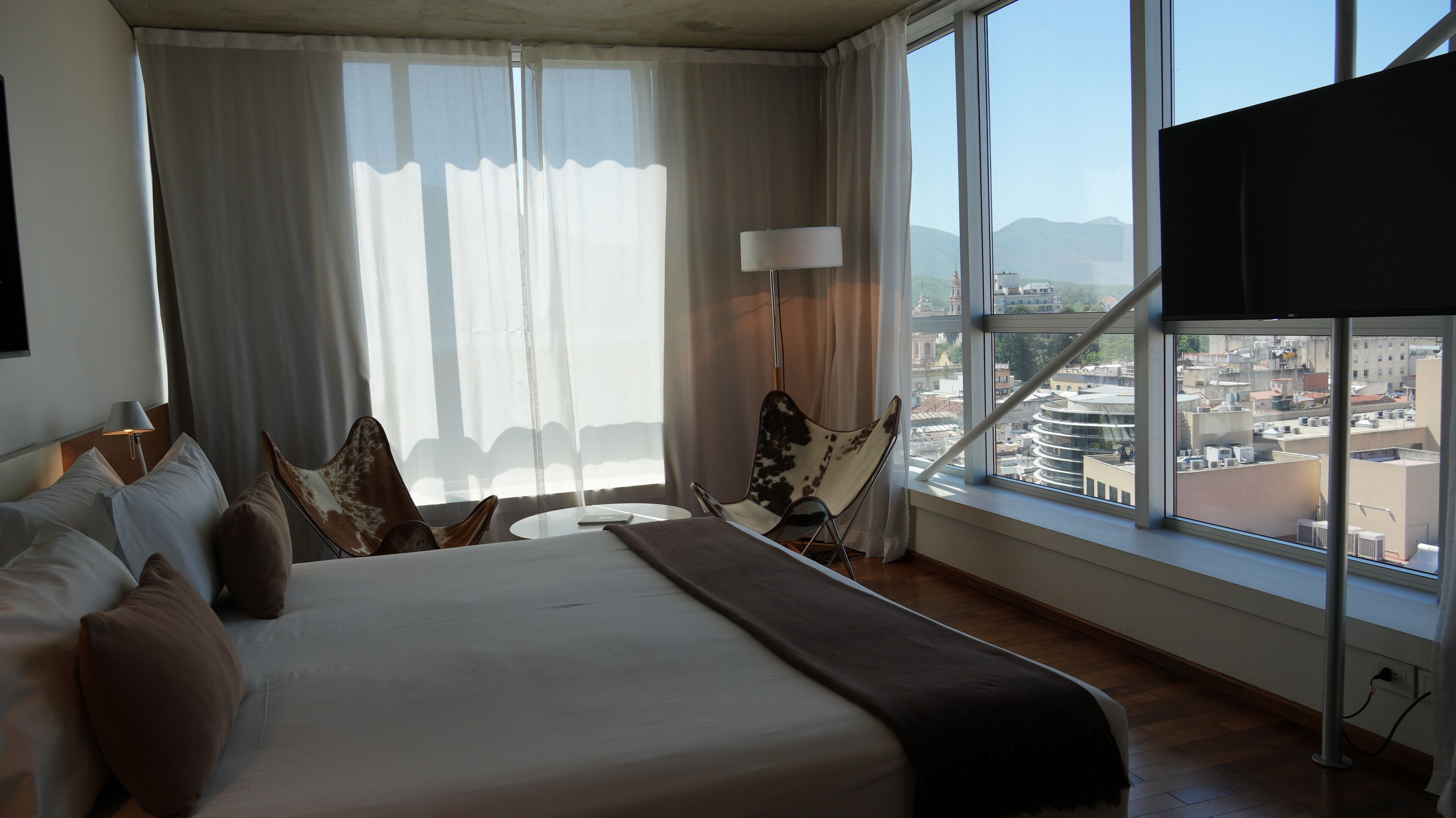 A hotel room with a large bed, a chair, a table, and a view of the city through large windows.