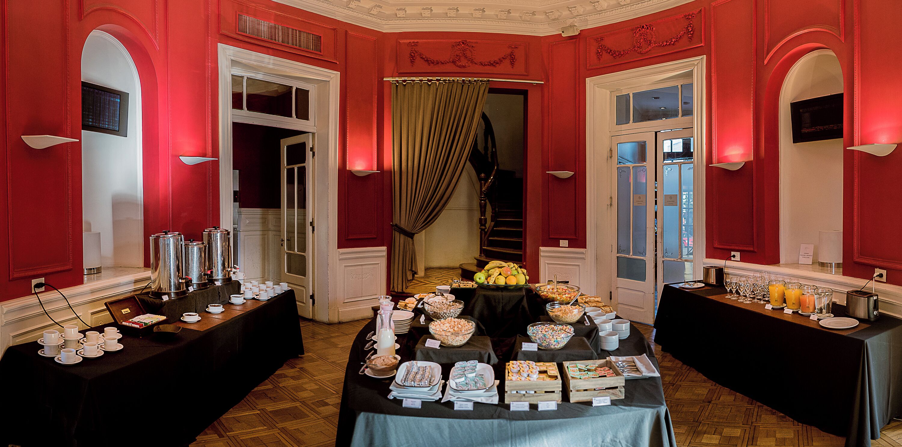 A room with red walls, a large table set with various food and drink items, and a view of a staircase leading to another room.