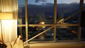 A room with a large window overlooking a cityscape at dusk, featuring a modern chair and a lamp.