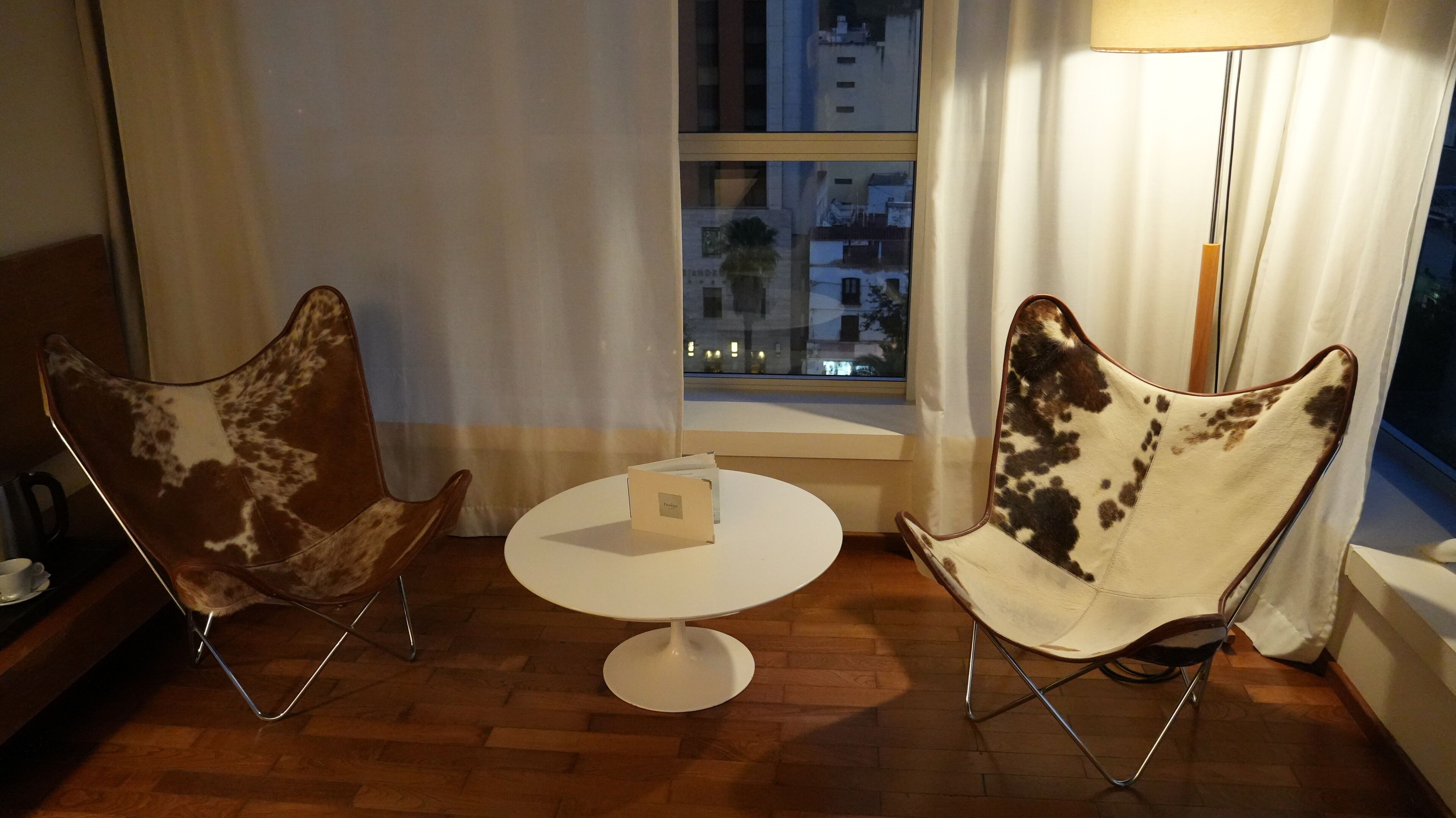 Deux chaises modernes au design original, une petite table ronde et une vue sur le paysage urbain, visible à travers une fenêtre ornée de rideaux.