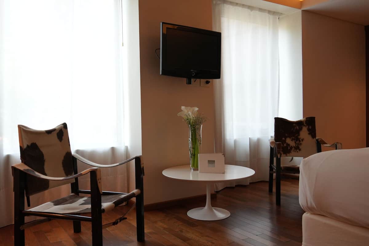 A hotel room with a bed, a TV mounted on the wall, a small round table with a vase of flowers, and two chairs with a unique design.