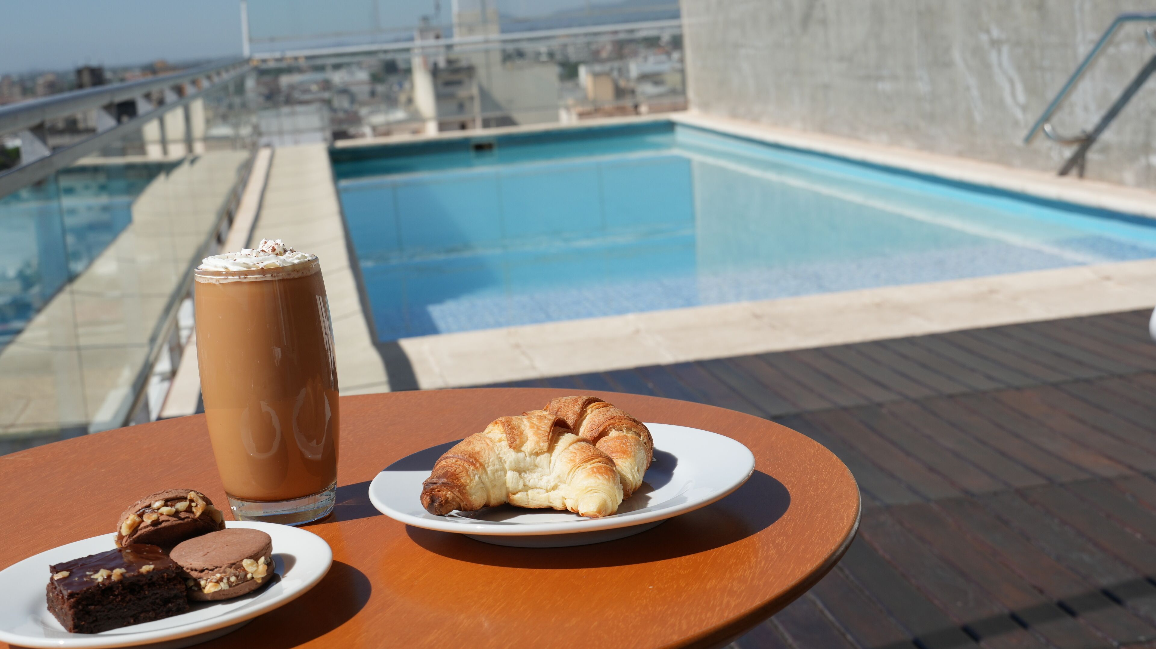 Una mesa con una bebida, un croissant y dos brownies en una terraza en la azotea con una piscina al fondo.