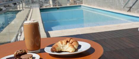 Una mesa con una bebida, un croissant y dos brownies en una terraza en la azotea con una piscina al fondo.