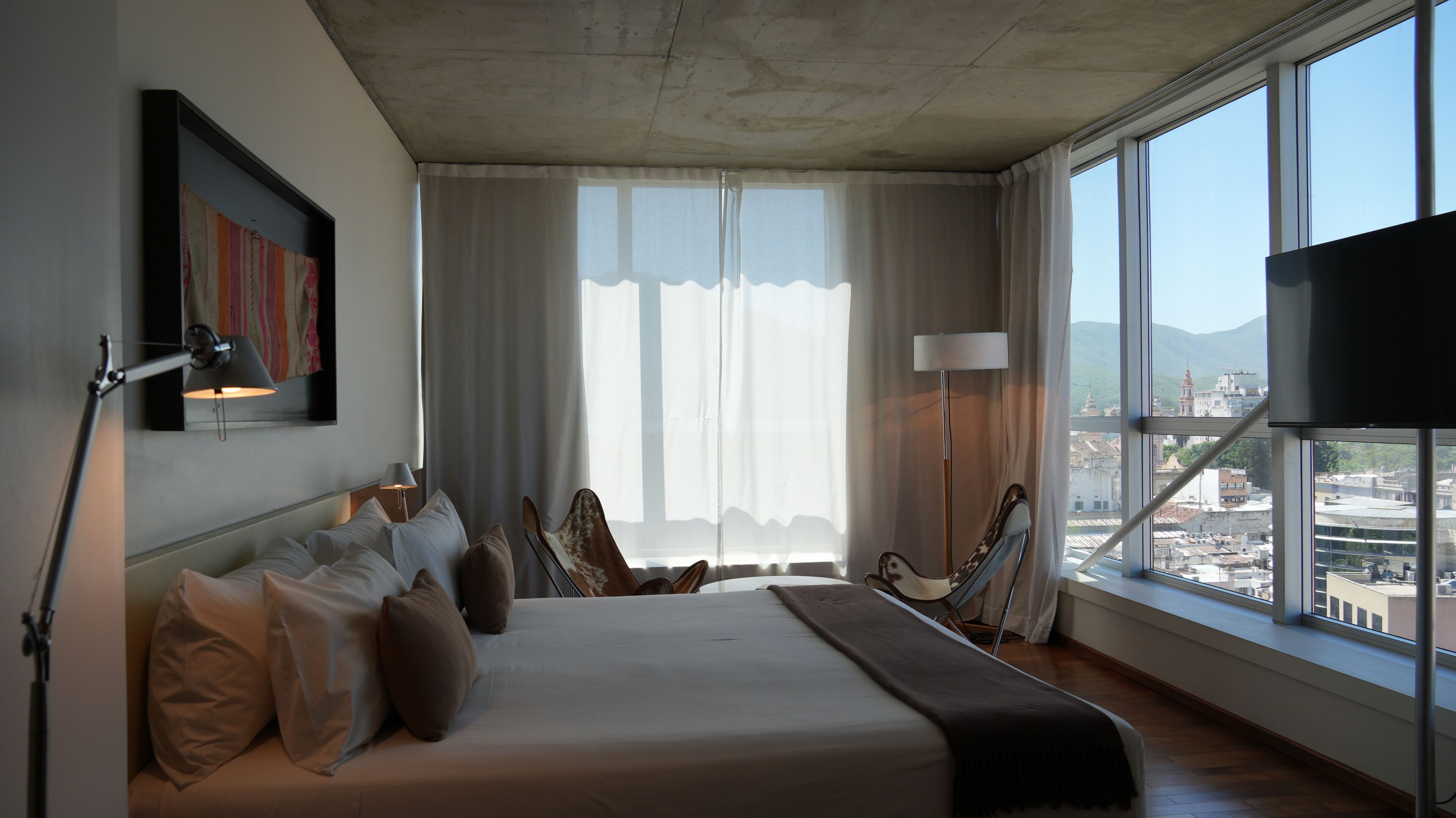 A modern hotel room with a large bed, multiple pillows, a desk lamp, a chair, and a view of the city through large windows.