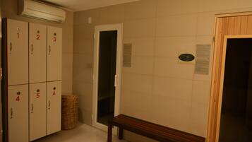 Habitación de hotel con taquillas numeradas, un banco, un cesto de basura y una puerta que da a otra habitación.