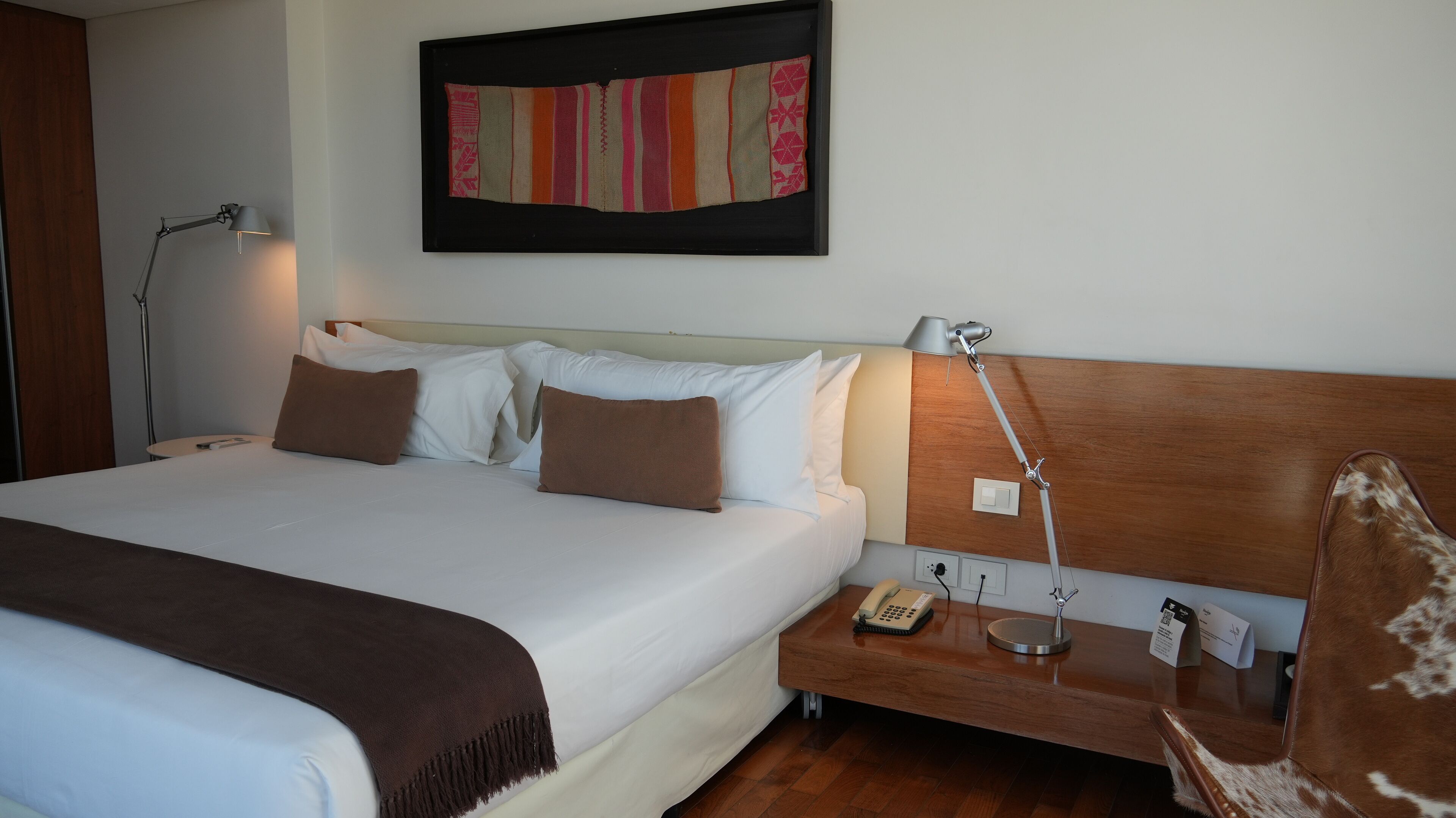 A neatly made bed with white and brown bedding, a wooden headboard, a bedside table with a lamp, a phone, and a small nightstand.