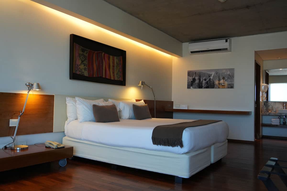 A modern hotel room with a large bed, bedside lamps, a wooden headboard, a desk with a telephone, and artwork on the walls.