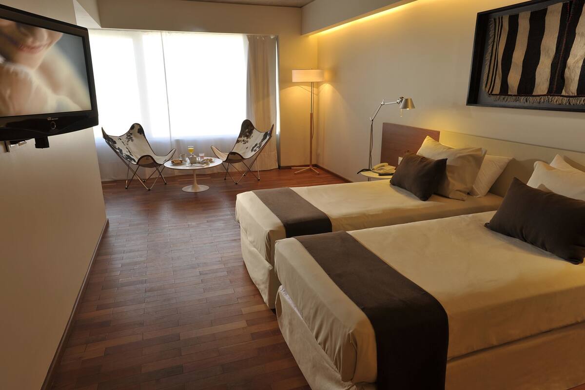 A hotel room with two beds, a TV, a desk with a lamp, a chair, and a window with curtains.