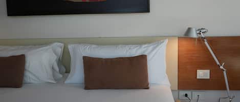 A hotel room with a bed, two pillows, a bedside lamp, a telephone, and a wall-mounted light switch.