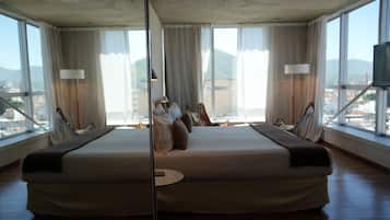 Un dormitorio con espejo, una cama grande, dos mesitas de noche y vistas a un paisaje urbano a través de amplios ventanales.