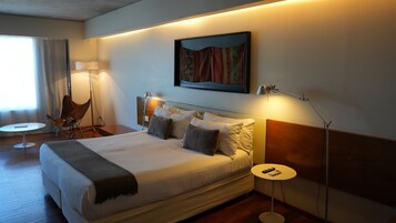 Habitación de hotel con una cama grande, lámparas de noche, una silla, una mesita y un cuadro en la pared.