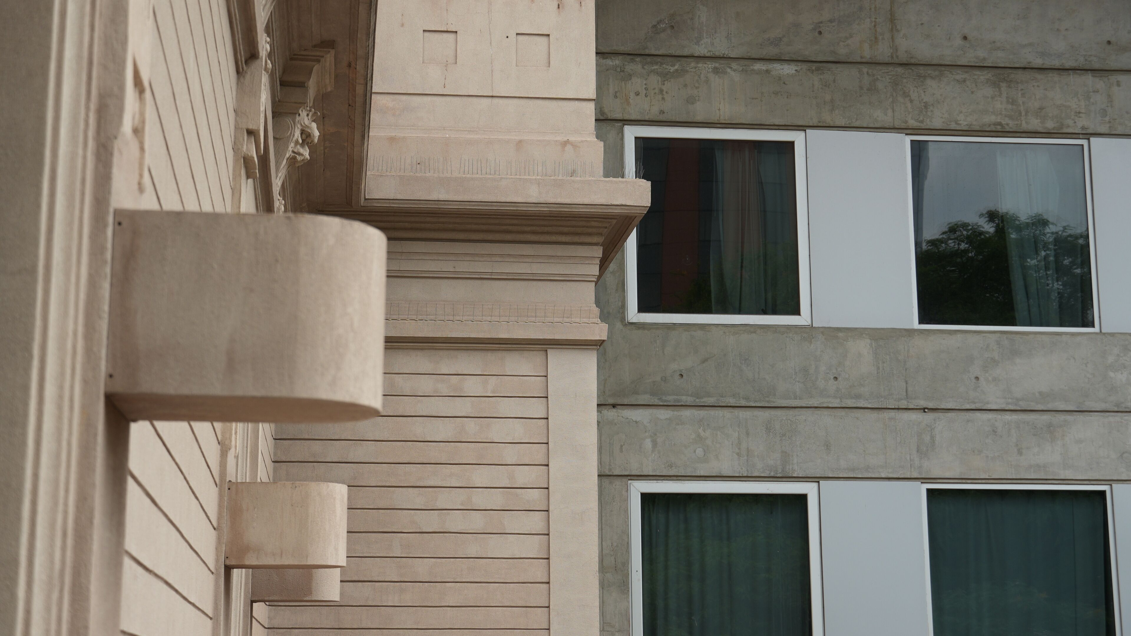Une façade d’immeuble de couleur beige, avec une partie en béton et trois fenêtres dotées de rideaux.