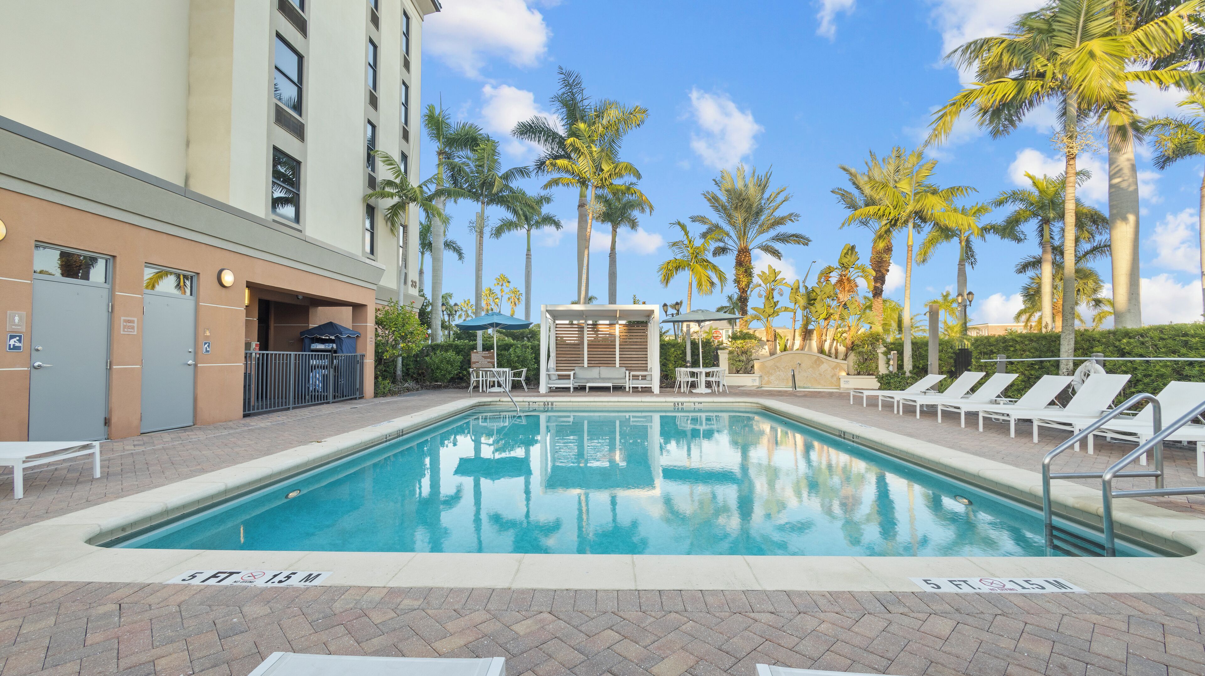 A swimming pool with lounge chairs and a covered seating area, surrounded by palm trees and a building with a beige facade.