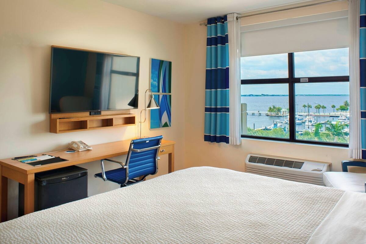 A hotel room with a bed, desk, chair, and a large window with a view of the marina.