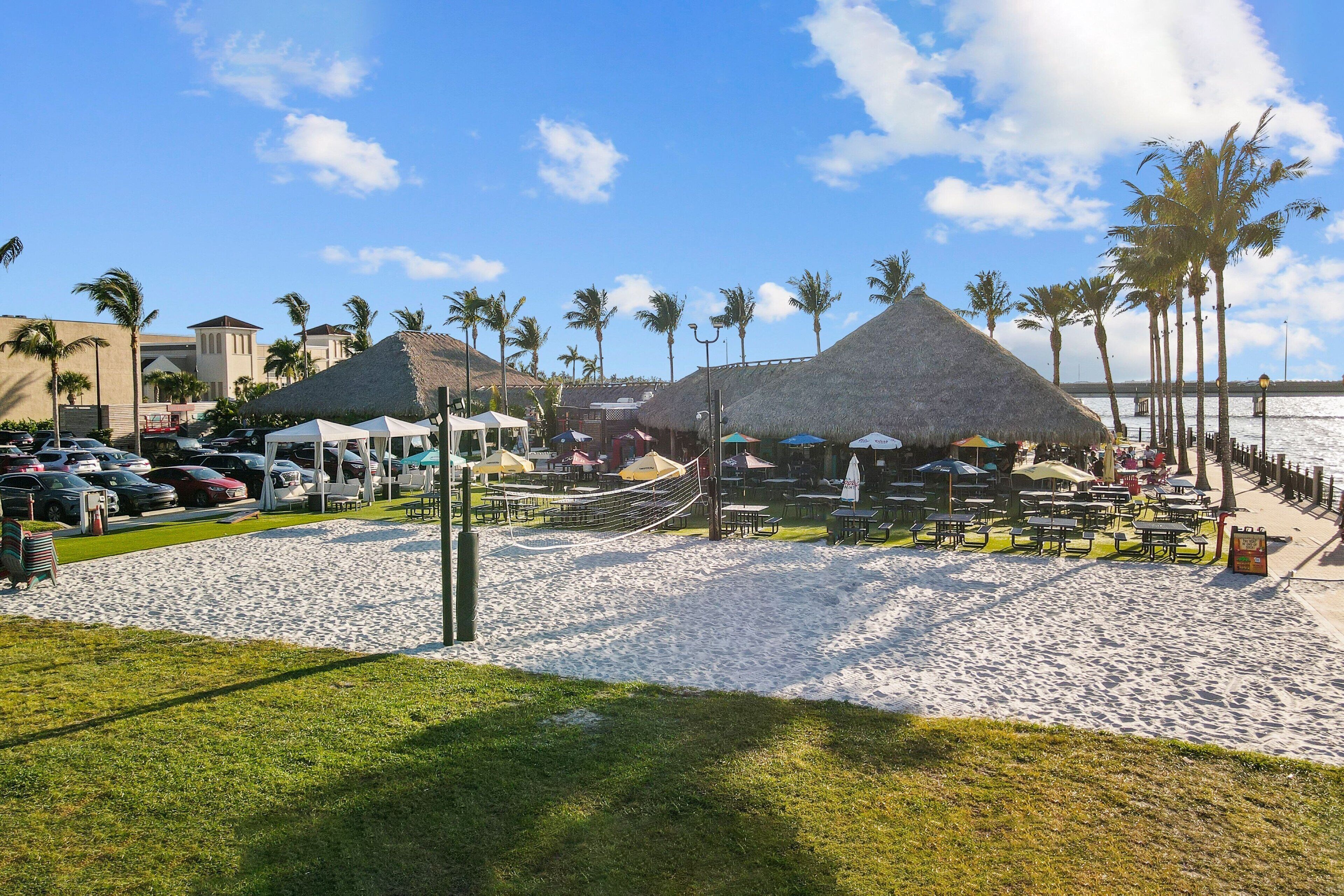 A beachside resort with thatched-roof structures, palm trees, and a sandy beach area with umbrellas and tables.