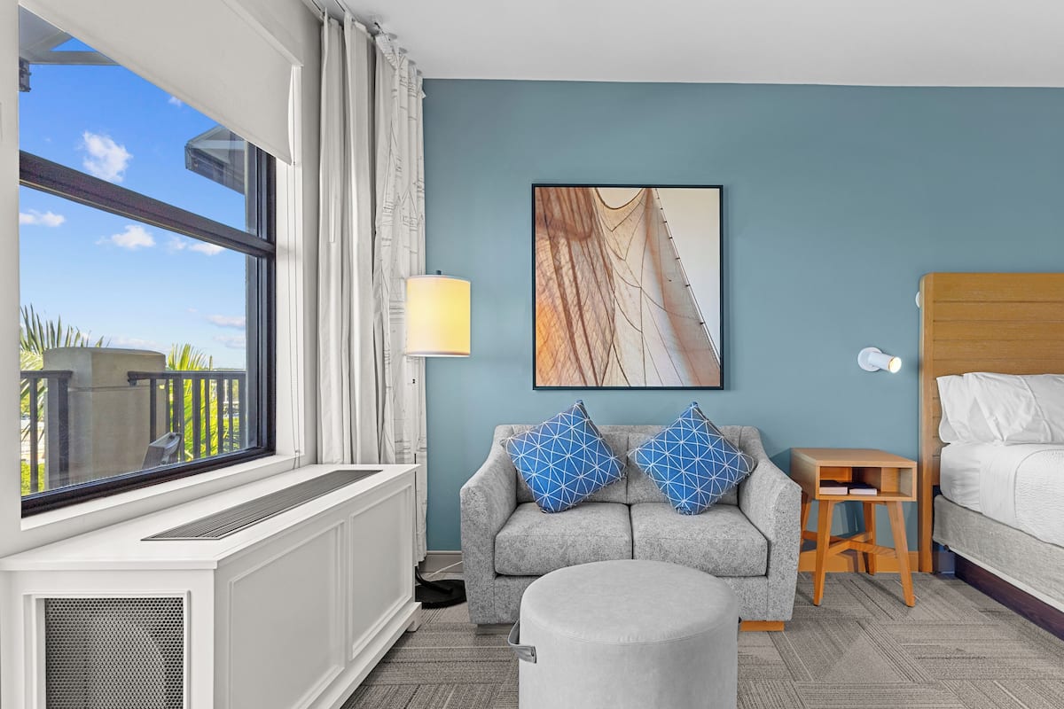 A hotel room with a bed, a grey sofa with blue patterned cushions, a small wooden nightstand, a wall-mounted lamp, a framed artwork, and a window with a view of the outdoors.