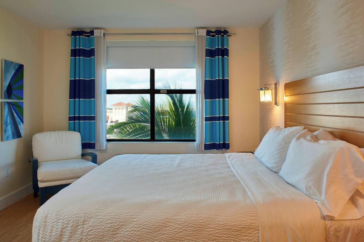 A hotel room with a large bed, a chair, a window with curtains, and a view of palm trees and buildings outside.