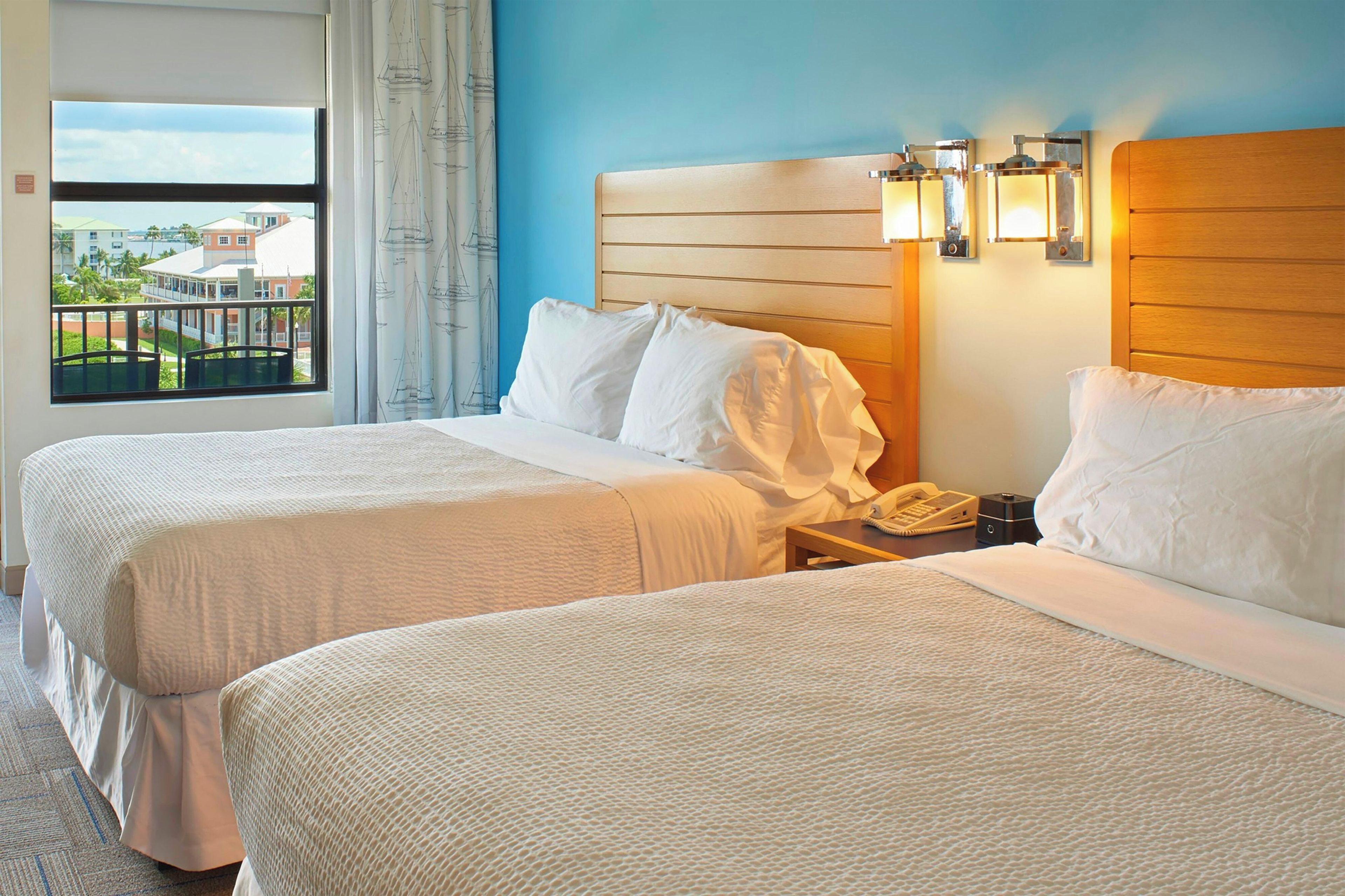 A hotel room with two beds, a wooden headboard, a nightstand with a phone, and a window with a view of buildings.