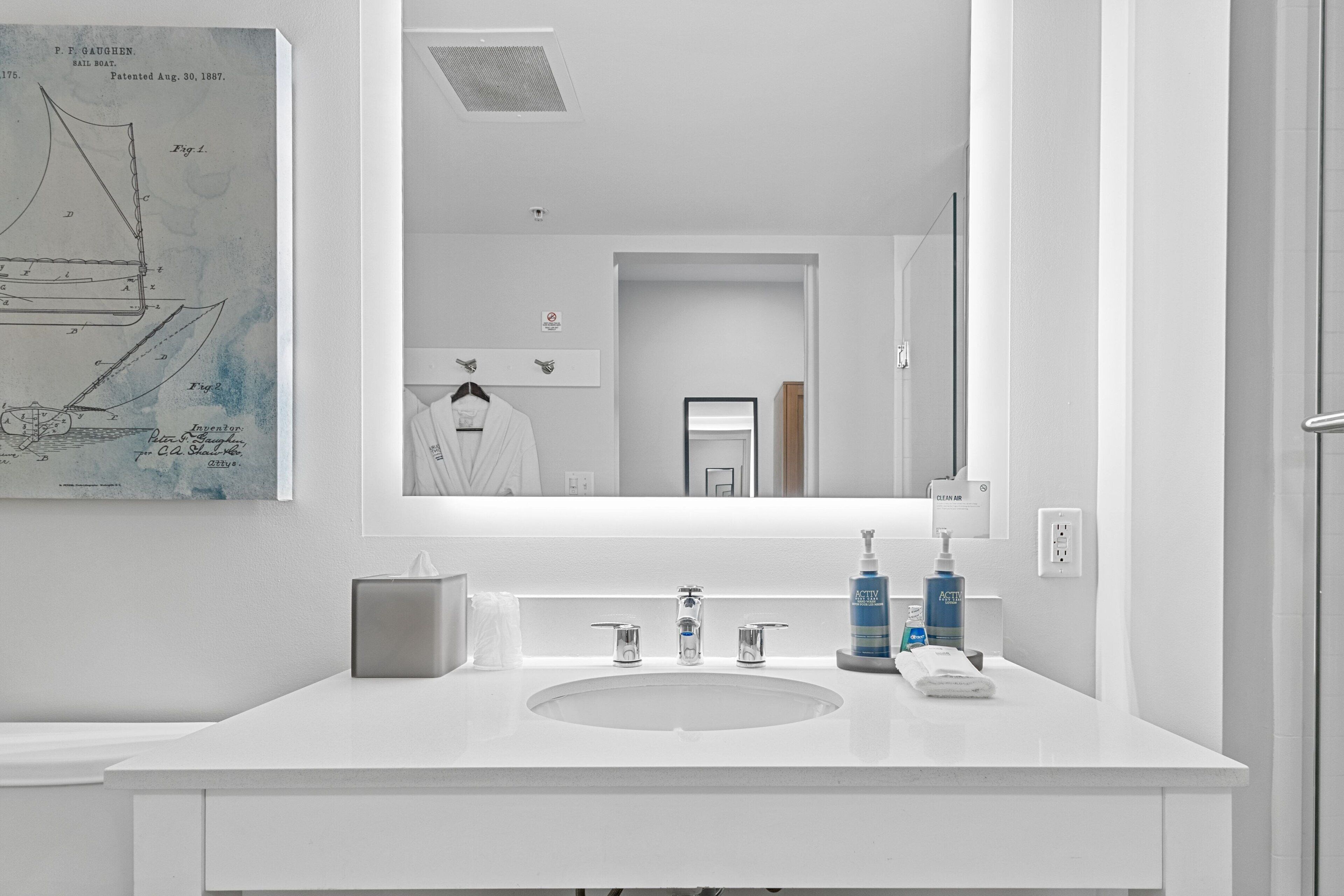 A modern bathroom with a large mirror, white sink, and two faucets. There is a framed nautical chart on the wall and a towel rack with a white towel.