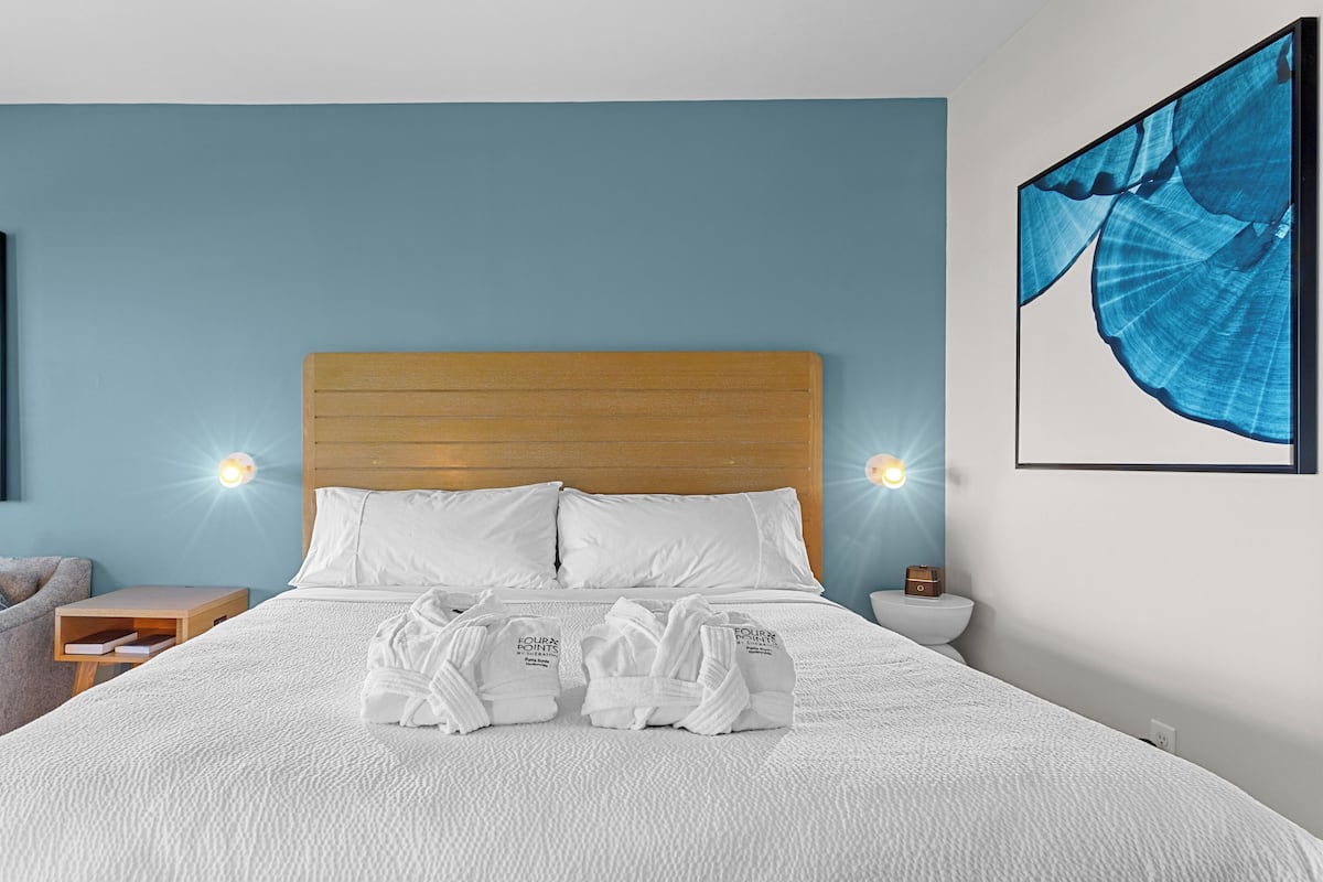 A neatly made bed with white linens, a wooden headboard, and two bedside lamps. There are two pillows, one with a blue geometric pattern. A pair of white bathrobes is folded on the bed.