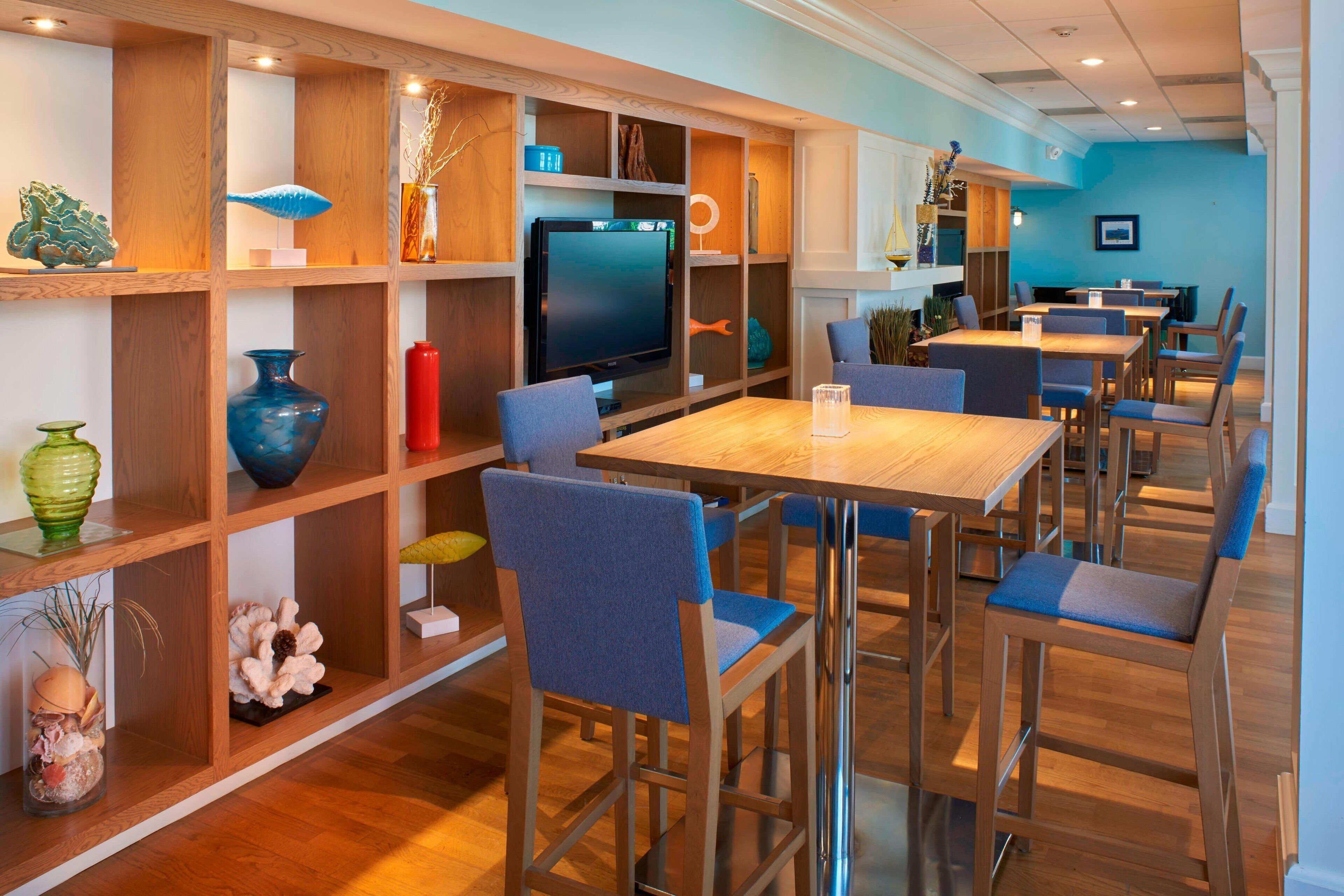 A modern dining area with wooden tables and blue upholstered chairs, a television, and decorative items on shelves.
