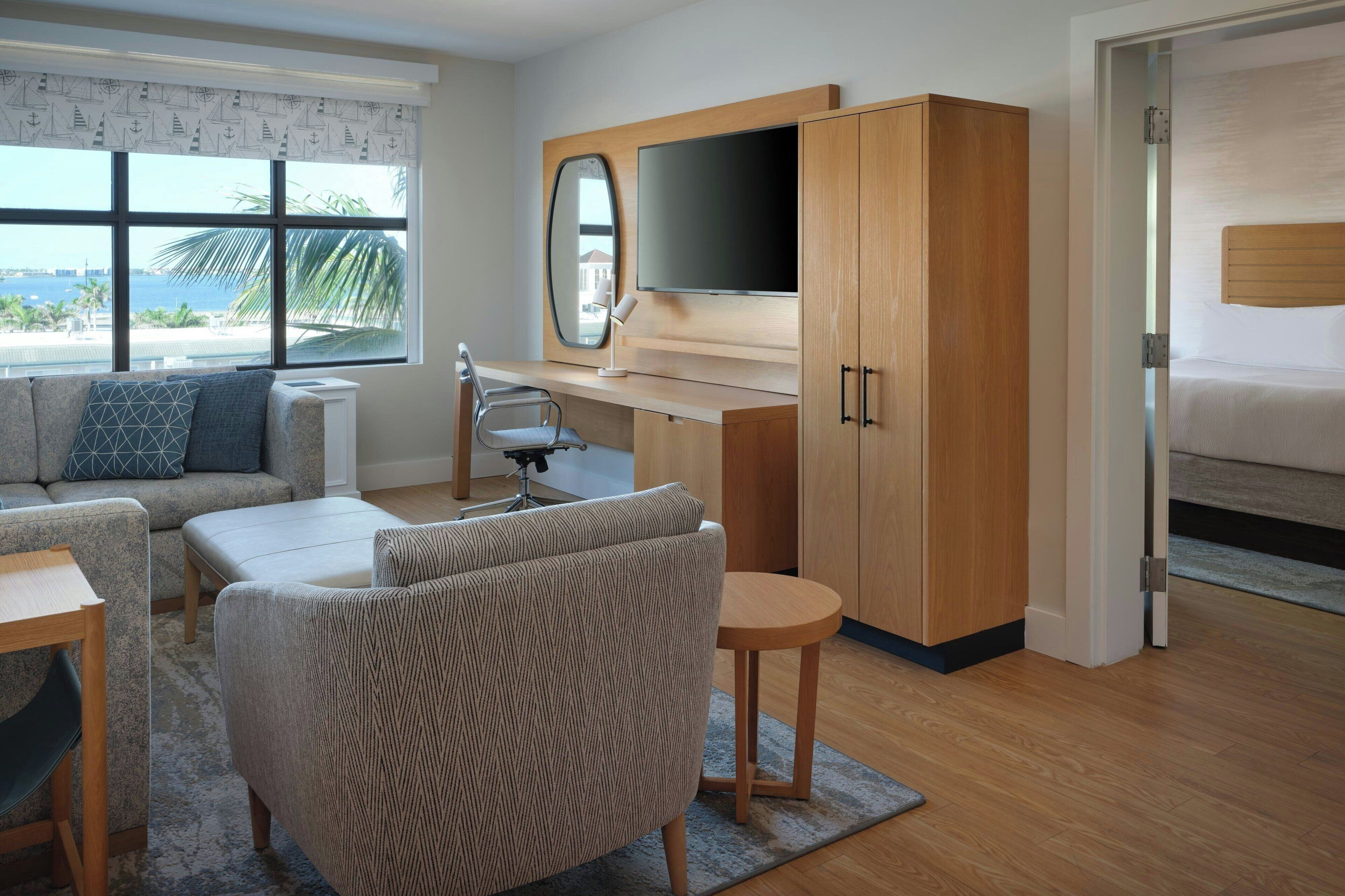 A modern hotel room with a large window, a bed, a desk with a TV, and a seating area with a sofa and chairs.