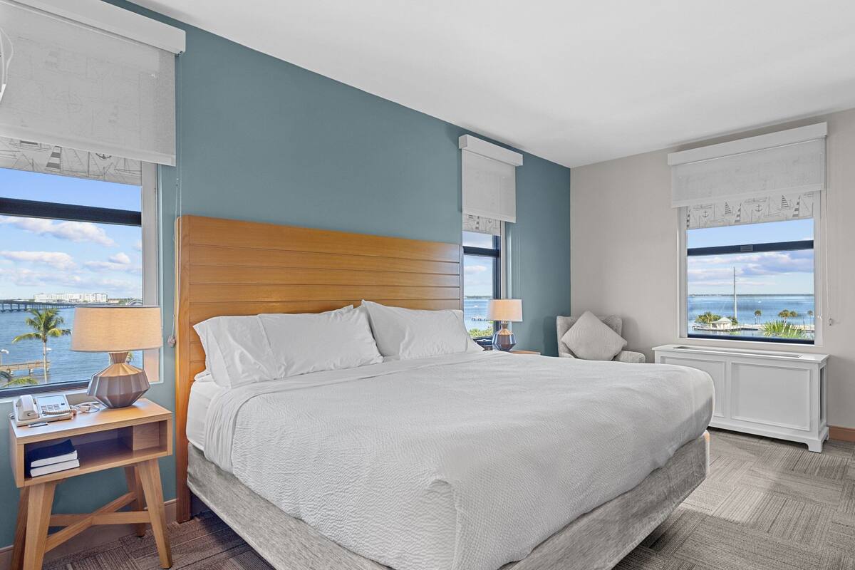 A hotel room with a large bed, wooden headboard, two bedside tables with lamps, a nightstand, and a view of the sea through large windows.