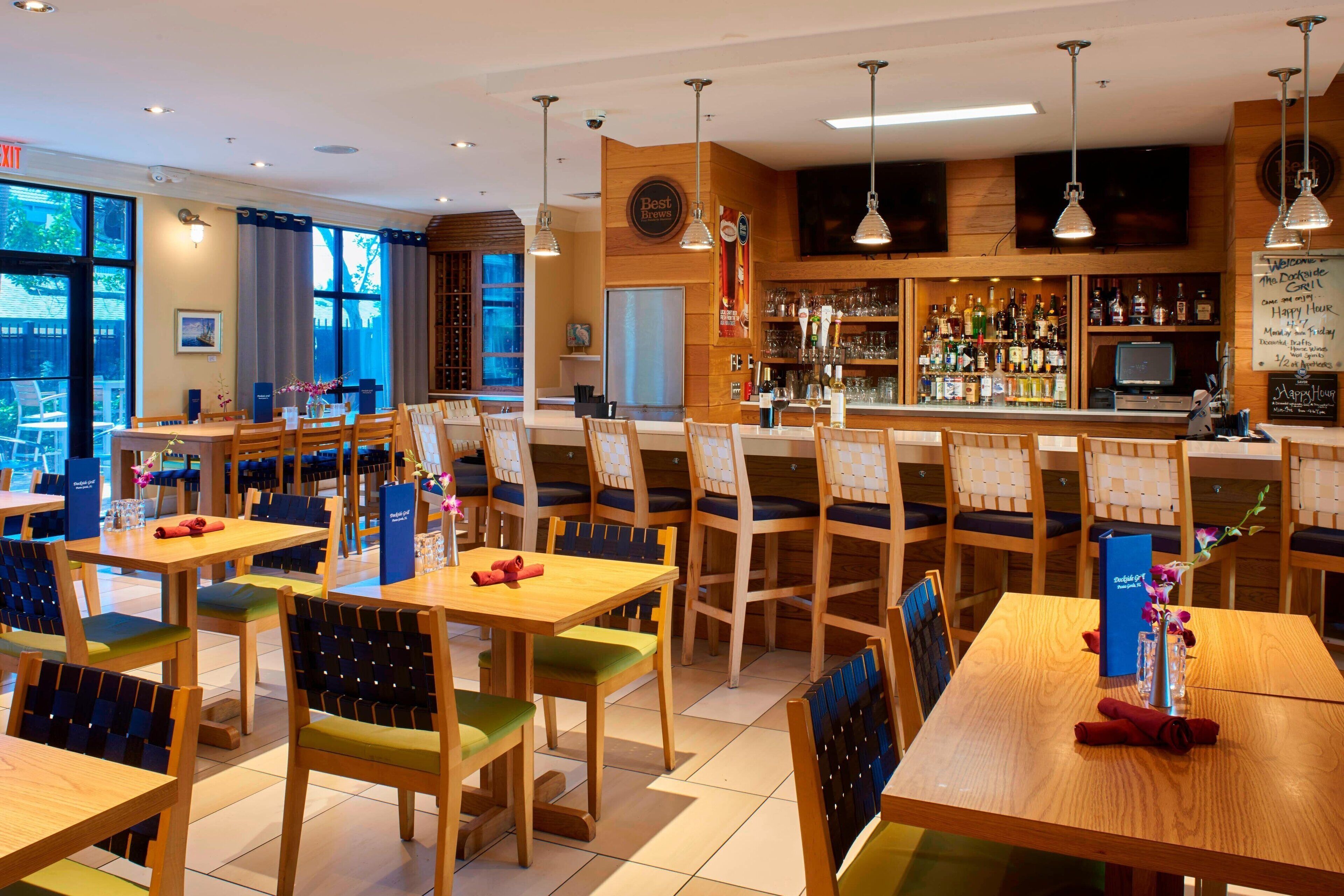 Un restaurante con mesas y sillas de madera, una zona de bar con varias botellas y vistas al exterior a través de grandes ventanales.