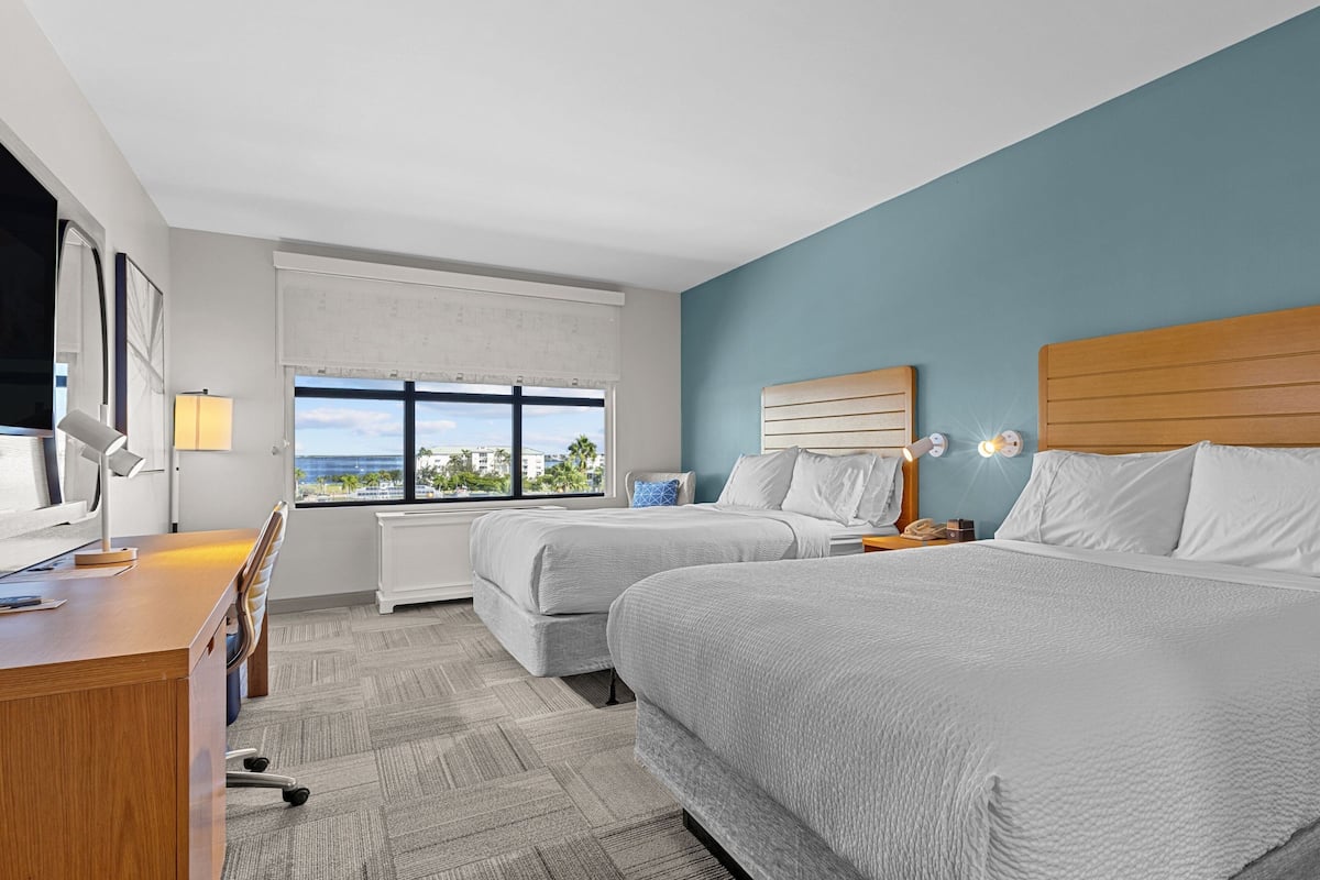 A hotel room with two beds, a desk, a lamp, and a view of the outdoors.