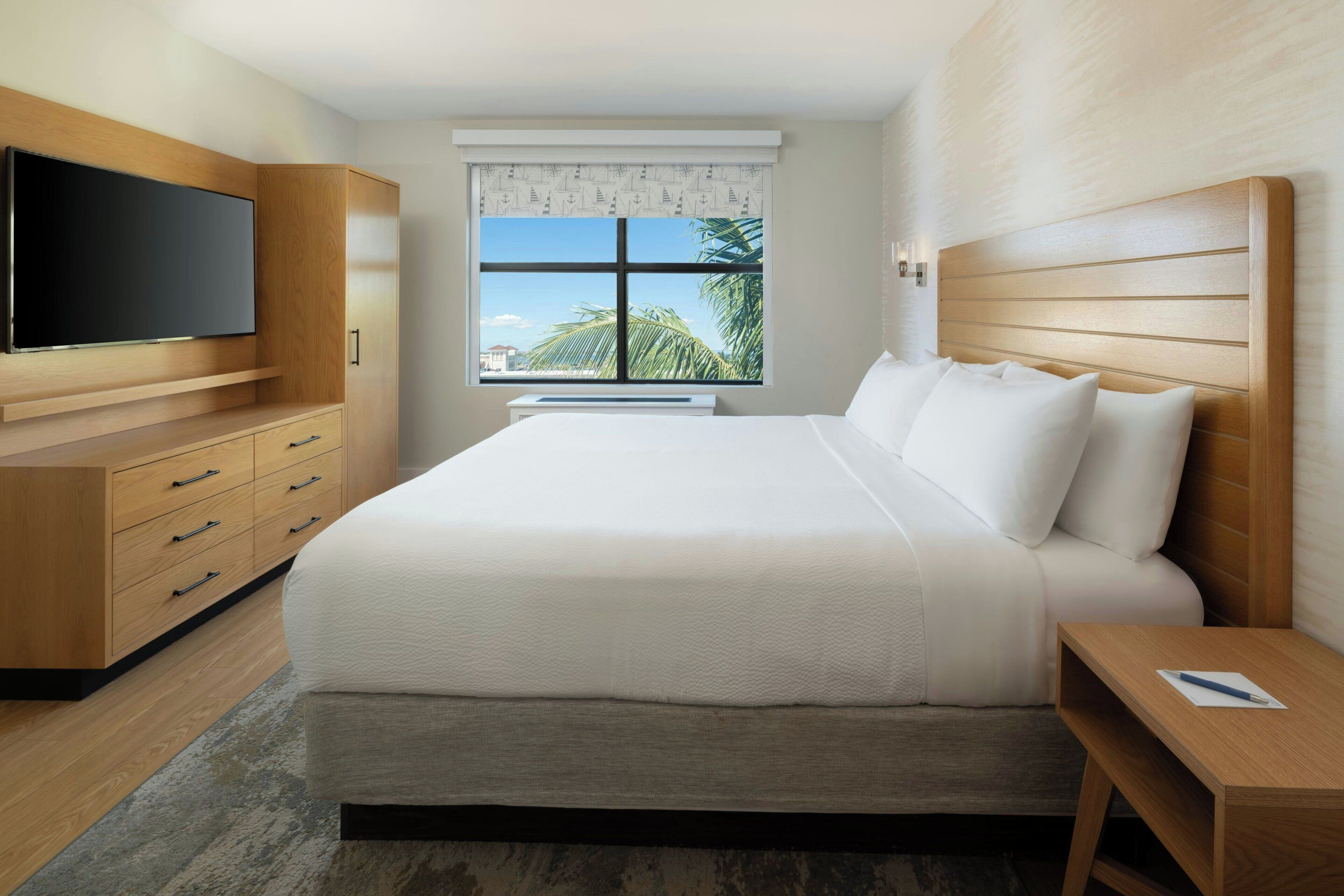 A modern hotel room with a large bed, wooden wardrobe, flat-screen TV, and a window with a view of palm trees.