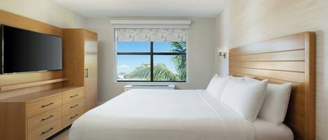 A modern hotel room with a large bed, wooden wardrobe, flat-screen TV, and a window with a view of palm trees.